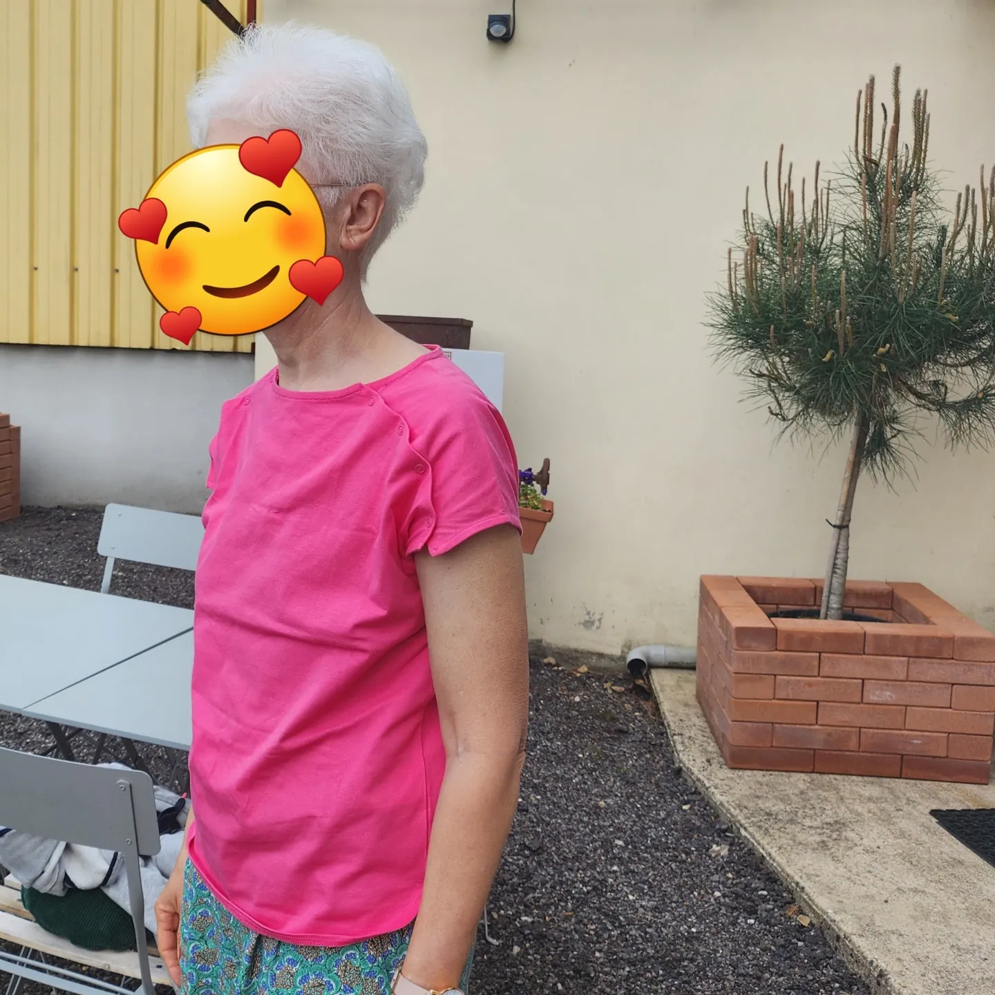 Personne aux cheveux courts argentés portant un tee-shirt rose, avec le visage masqué.