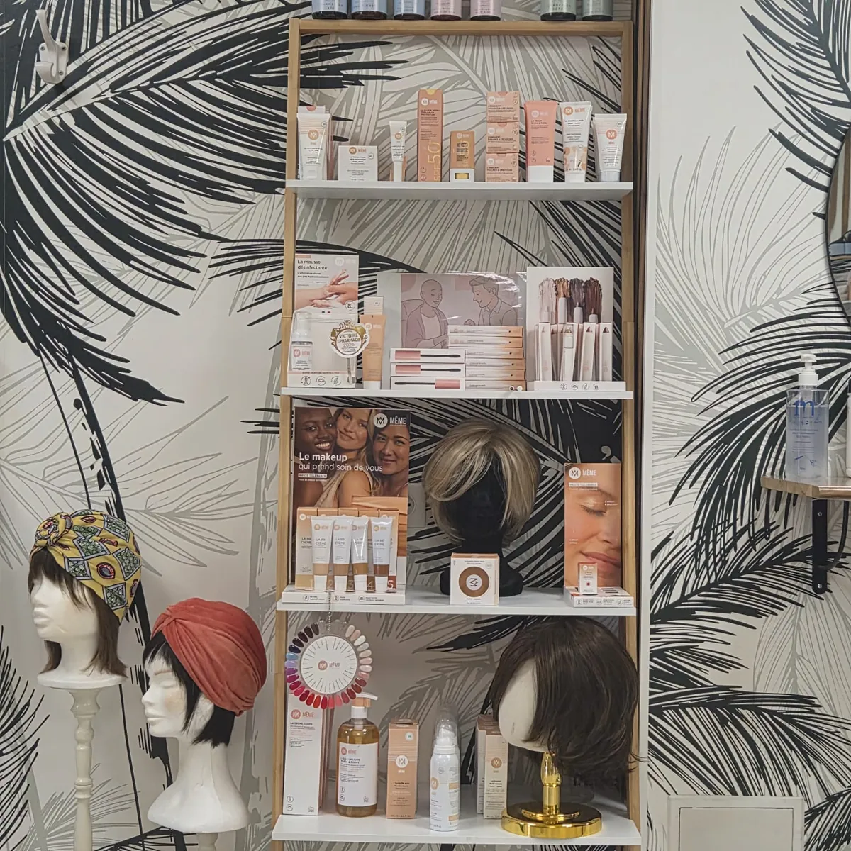 Présentoir de soins et de maquillage MÊME, entouré de perruques et de turbans aux Ateliers d’Eve&Marco.